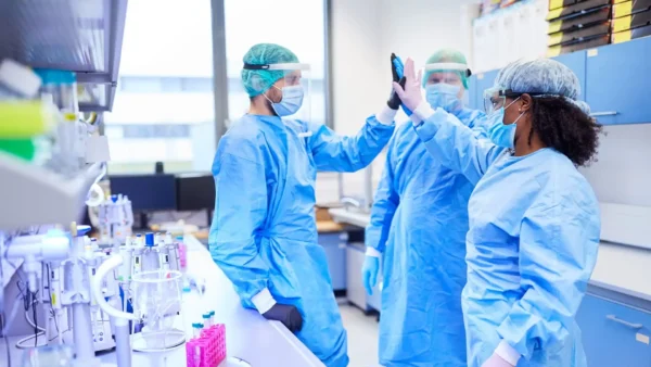 Three scientists in full PPE, including gowns, masks, and face shields, give each other high-fives in a laboratory.