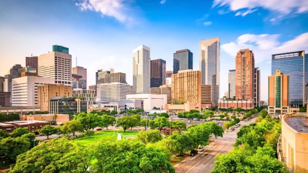 A panoramic view of a modern city skyline with numerous high-rise buildings and skyscrapers under a bright, partly cloudy blue sky. Lush green trees line the foreground streets.